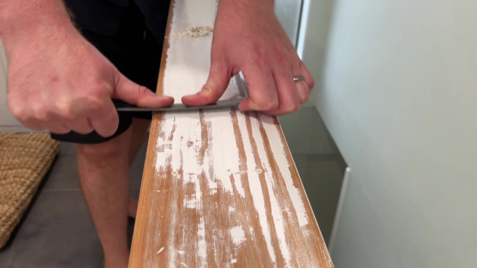 一个男人的手用一把钢尺刮掉木栏杆顶部的白色油漆的特写