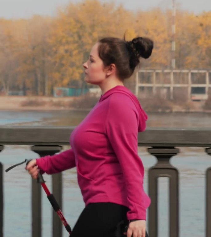 一个身材魁梧的女人拿着北欧的杆子沿着河岸走着。垂直视频