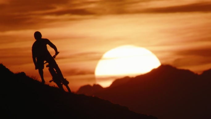 在日落期间，确定的剪影山地自行车手在戏剧性的天空下骑自行车下山的锁定镜头