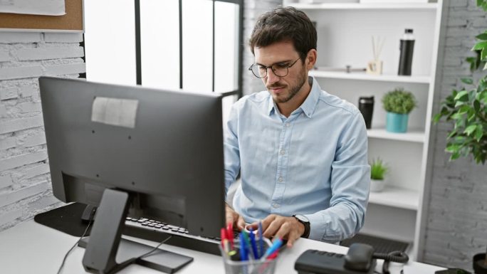戴眼镜的西班牙裔男子在一间现代化的办公室里工作和庆祝，那里有电脑和办公用品