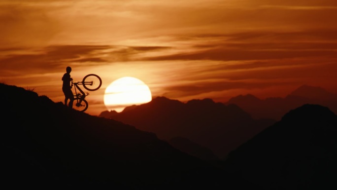SLO MO锁定的剪影山地自行车手携带和推动自行车，而移动上山对抗戏剧性的橙色天空