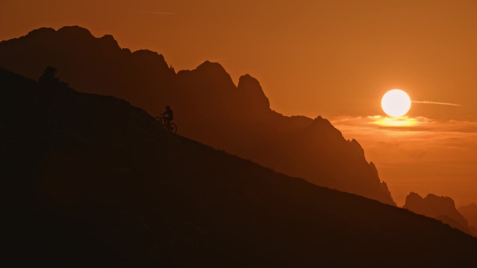 慢动作低角度远镜头的剪影运动员骑山地车上山对橙色的天空在日落