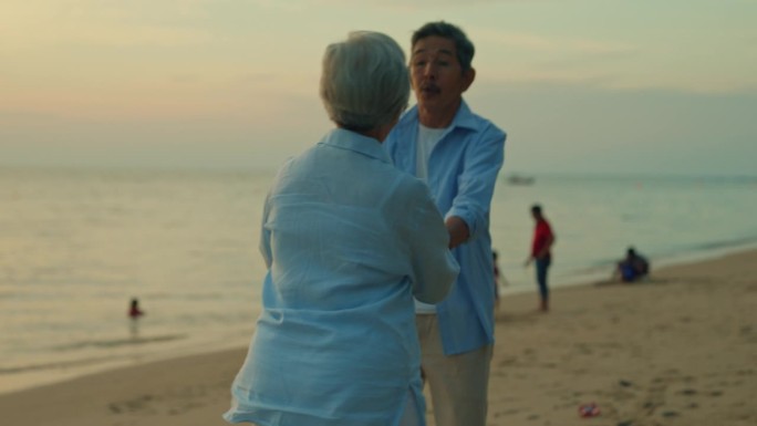 欢快的亚洲老夫妇手牵着手在沙滩上跳舞，欣赏夕阳下的大海。一个快乐的老年人享受退休后的旅行生活方式。
