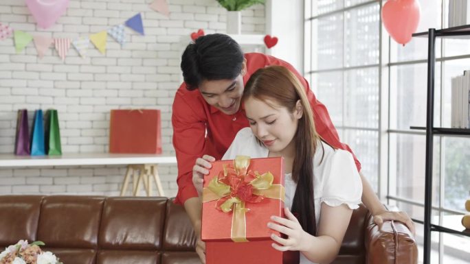 微笑的年轻亚洲丈夫或男友闭上眼睛看着他的妻子或女友，在客厅里送礼盒。生日惊喜，情人节和周年纪念，在家
