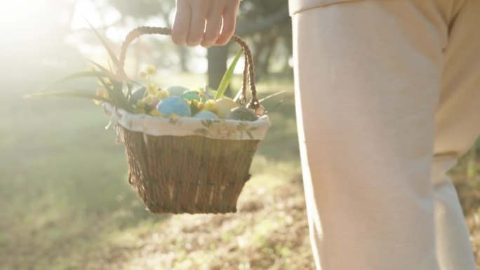 一个穿白衣的女人提着一篮子复活节彩蛋在森林里散步，朝着阳光走去。林地环境中的电影照明。
