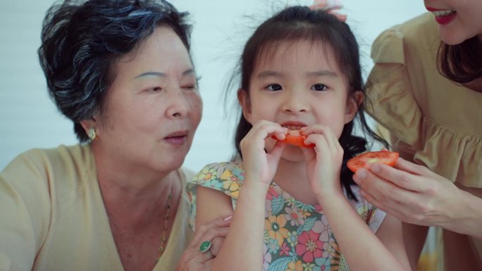 专注于美丽的亚洲母亲喂西红柿给她的小可爱的女儿，她站在她的妈妈和奶奶在家里的厨房。开心的奶奶看着她的