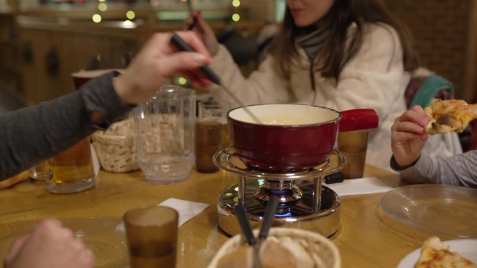 人们在餐馆里吃奶酪火锅，特写是用手把一块面包蘸进融化的奶酪里，红色的锅放在加热器上