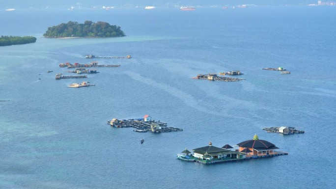 小船驶过漂浮的网笼和漂浮的清真寺，它们停泊在岸边平静的蓝绿色水域上，远处是热带小岛、渡轮和油轮