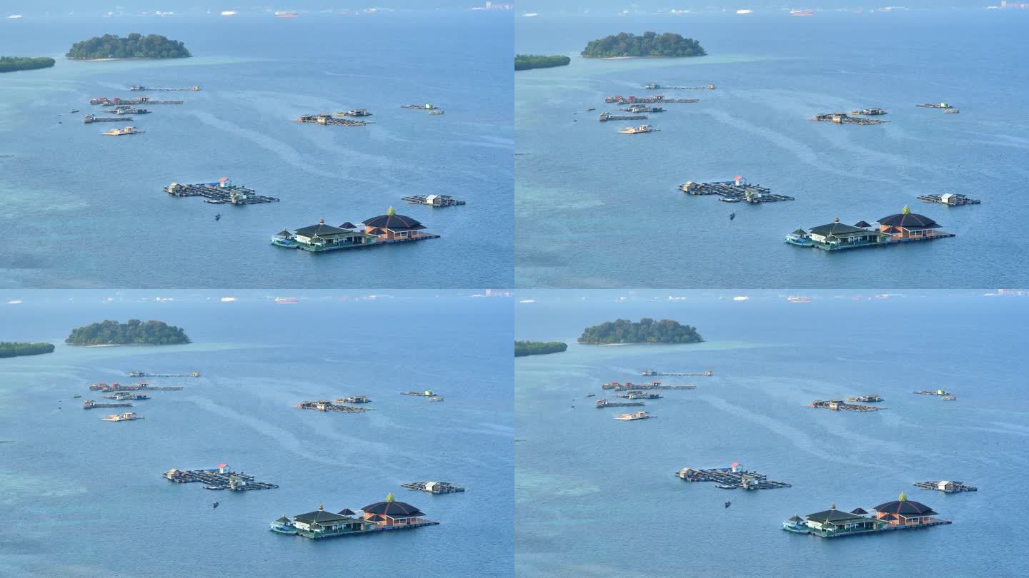 小船驶过漂浮的网笼和漂浮的清真寺，它们停泊在岸边平静的蓝绿色水域上，远处是热带小岛、渡轮和油轮