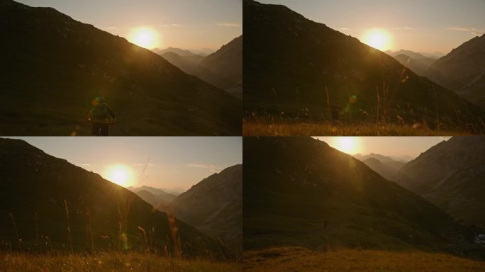 男子骑自行车在草地上对雄伟的山脉在明亮的日落的后视图