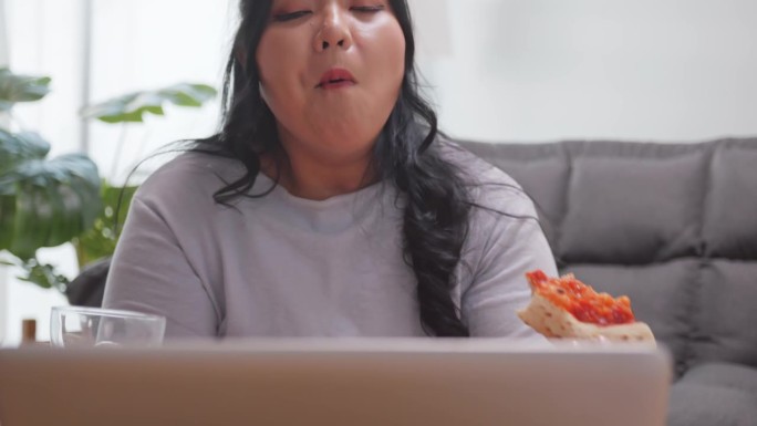 一个胖乎乎的亚洲女人躺在家里的沙发上享受着披萨和软饮料。