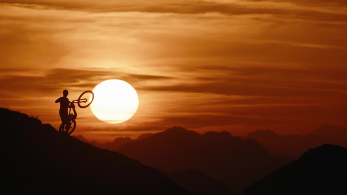 SLO MO锁定的剪影山地自行车手携带和推动自行车，而移动上山对戏剧性的日落天空
