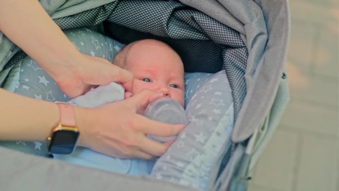 一名新生婴儿躺在婴儿车里，母亲用奶瓶喂初学走路的女婴