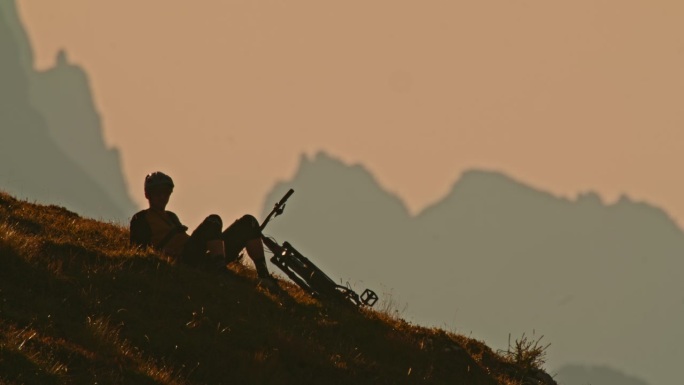 SLO MO锁定镜头，放松的男性山地自行车手斜倚在山上对着晴朗的天空在日落