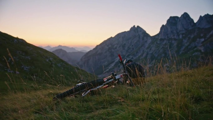 手持拍摄的男子骑自行车坐在草地上对落基山脉日落