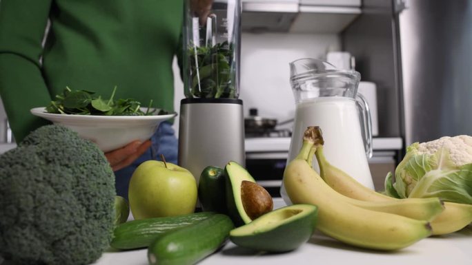 一名妇女把新鲜的绿色菠菜放在搅拌机里，在家里的厨房里用蔬菜和水果制作健康的排毒奶昔