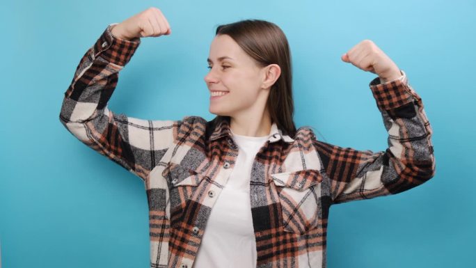强壮的健身年轻女子穿着休闲衬衫，展示双手二头肌，展示力量，在工作室淡蓝色背景墙前单独摆姿势。人们的生