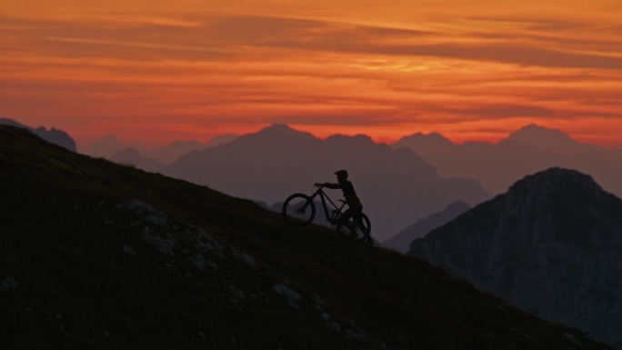 SLO MO手持镜头的轮廓男性运动员推动山地自行车上坡对戏剧性的橙色天空