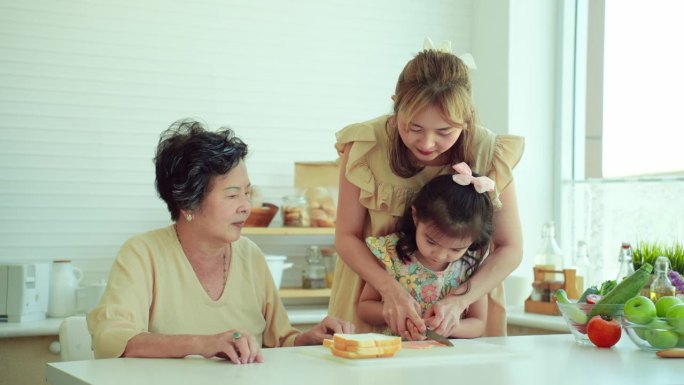 美丽的亚洲母亲教她可爱的小6岁的女儿用菜刀切火腿片，准备三明治作为早餐在家里和奶奶。孙女喜欢学习如何