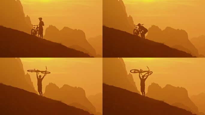 骑山地车的人扛着自行车站在山上，面对雾山和橙色天空的剪影