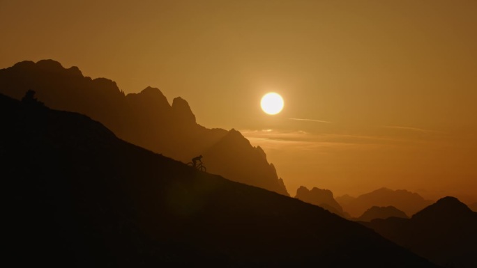 SLO MO低角度远拍的剪影骑自行车在山上对着橙色的天空日落