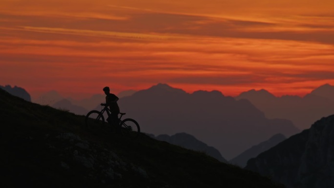 SLO MO手持拍摄的剪影男性骑自行车推山地车，而爬坡对戏剧性的橙色天空