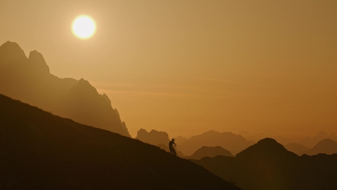 在雾蒙蒙的山脉和壮观的橙色天空下，骑自行车的人的剪影在山上的锁定镜头