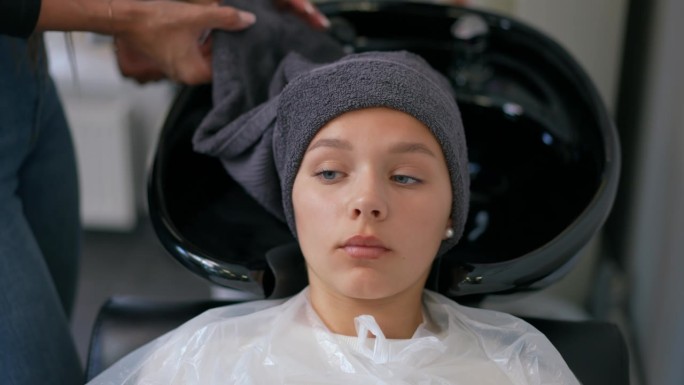 一位女发型师示范如何正确地将毛巾缠在顾客头上。一位女顾客躺在带水槽的美发师椅上。
