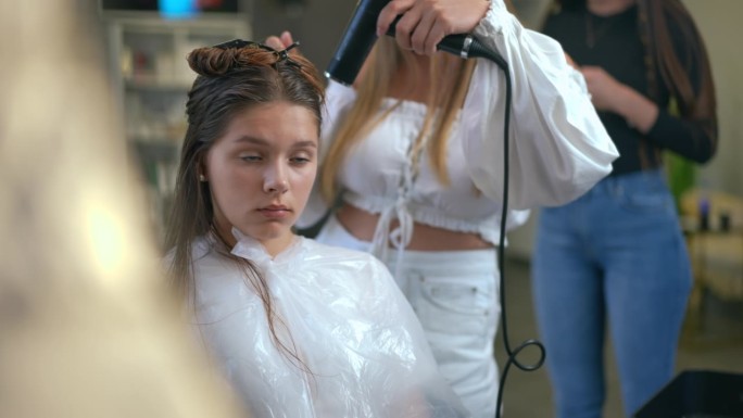 一个女孩坐在美发沙龙里看着镜子。女理发师用吹风机吹干女孩的湿头发。
