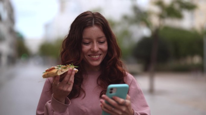年轻的红发女人边走边看她的智能手机，在街上吃着美味的披萨