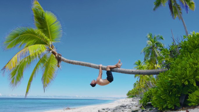 一个赤裸上身的男人在沙滩上放松，爬上一棵椰子树。天堂海滩上的瘦子