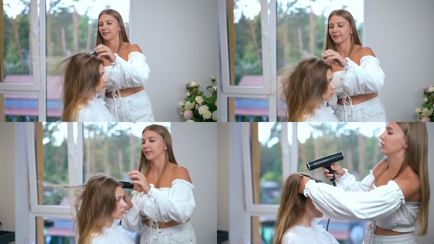 缓慢的运动。一位女理发师站在现代沙龙的窗户背景上，用吹风机吹干一个女孩的头发。