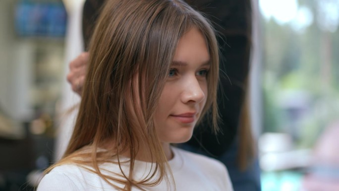 一个女孩坐在一家现代理发店里，一个女理发师站在后面，检查女孩头上的一缕头发的强度。