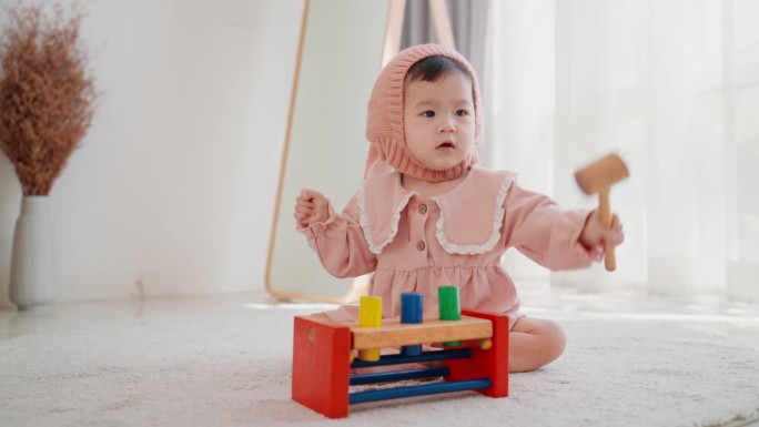 可爱的亚洲女婴坐在毛绒地毯上，穿着粉红色的衣服和帽子，在家里玩着五颜六色的木锤块玩具。玩一个充满活力