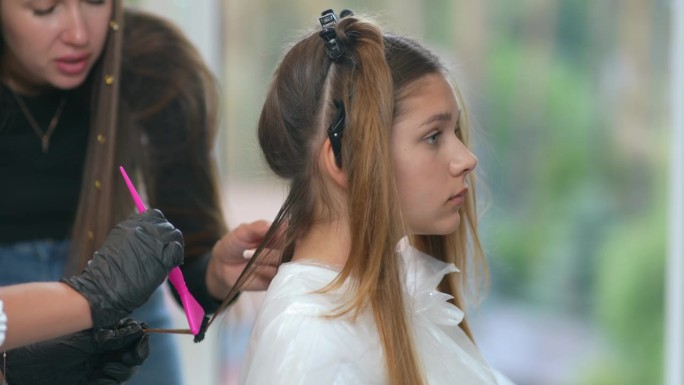 一位女发型师老师看着一位戴着黑色橡胶手套的新手发型师用梳子从客户头上梳下一缕头发。