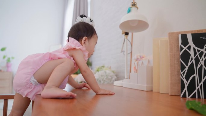 可爱的亚洲女婴勇敢地爬上妈妈的工作台，去够家里客厅里的东西。