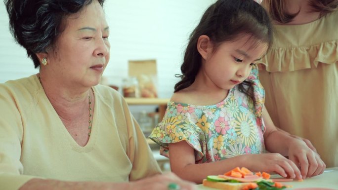 亚洲中年母亲和老祖母教他们的小女儿用刀切新鲜的胡萝卜和火腿，为家里的早餐准备一个健康的三明治。三代同