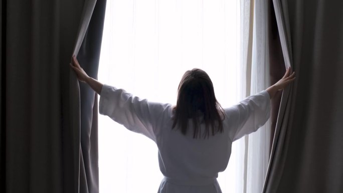 身穿白色浴袍的年轻女子站在窗前，用手拉开窗帘(特写，后景)。