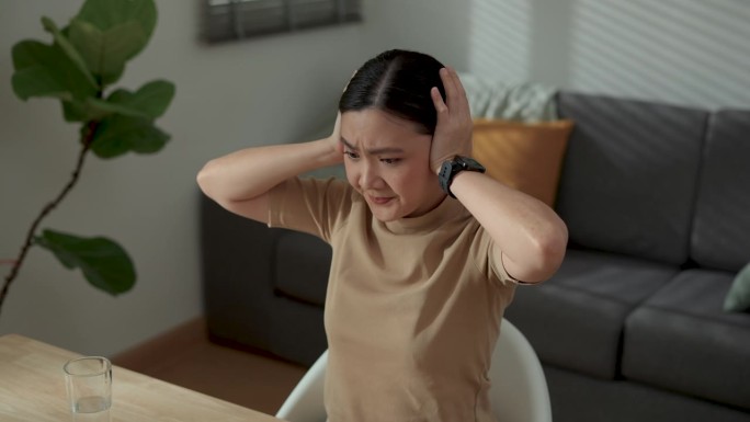 在家庭办公室使用笔记本电脑工作的亚洲女性感到无聊和厌烦，用手捂住耳朵。