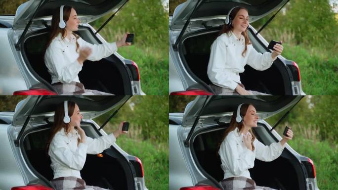一名年轻女子坐在一辆自然的汽车后备箱里，用智能手机通过视频连接说话