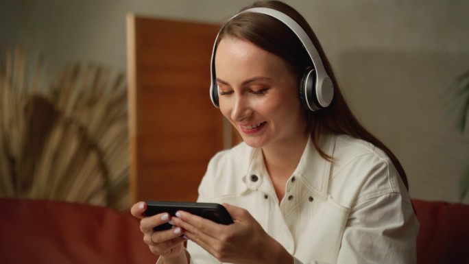 兴奋的年轻女子戴着耳机，在智能手机上玩在线游戏，网络直播。