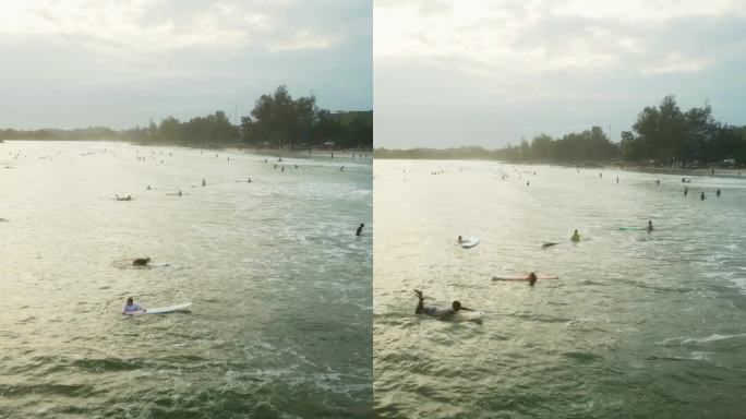 日落时海浪中的冲浪者鸟瞰图。密集的人群冲浪，在板上划桨，等待着海浪。充满活力的海景，在黄金时间从上面