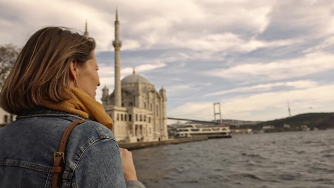 手持拍摄的女游客看着Ortakoy清真寺和博斯普鲁斯大桥在海上多云的天空