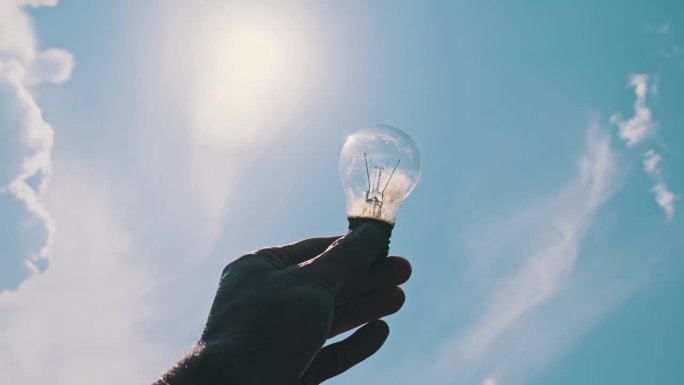 男性手拿白炽灯泡顶着明亮的太阳在蓝天