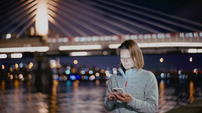 蓝色时刻，一名女子在博斯普鲁斯海峡边散步，用手机，背景是金角地铁大桥。