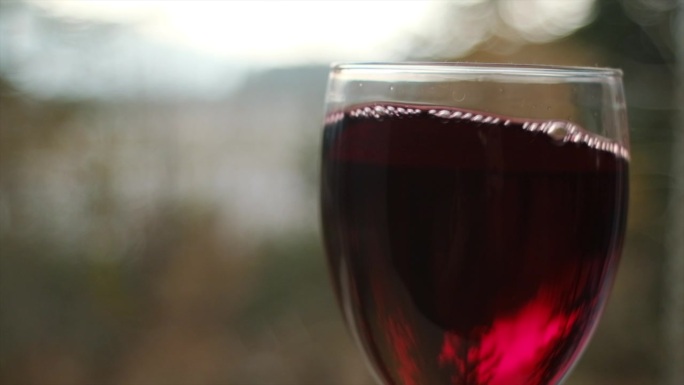 在自然的背景下，在阳光下摇曳着酒杯中的红酒。美丽的葡萄酒商业素材。高脚杯内的调酒过程特写。慢动作