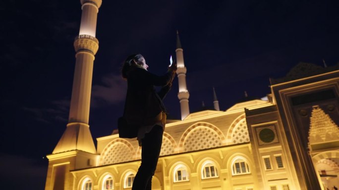 土耳其伊斯坦布尔，女游客用智能手机拍摄被灯光照亮的Çaml¹ca共和国清真寺的低角度手持照片