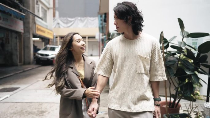 一对年轻的亚裔华人夫妇手牵着手，微笑着走在香港当地的城市街道上，享受着城市生活。爱情和约会概念