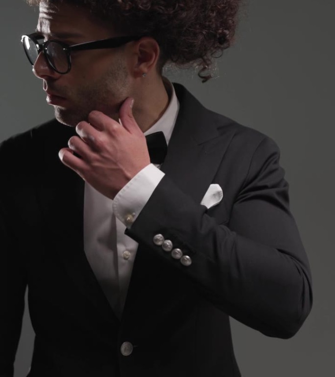 项目视频，年轻优雅的男子戴眼镜，穿着黑色燕尾服，触摸脸，在灰色背景前摆姿势