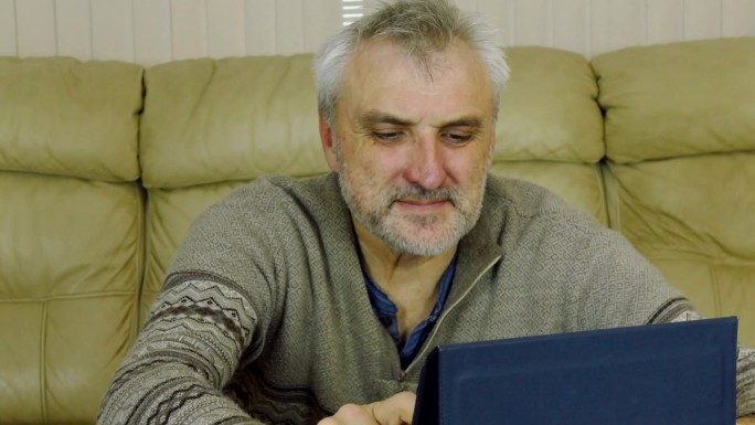 没刮胡子，头发花白的男人坐在家里的沙发上用平板电脑工作